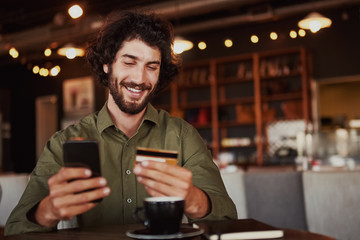 Happy caucasian bearded man holding mobile phone typing card data to make online payment sitting in...