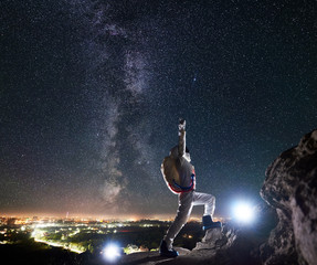 Side view of spaceman raising arms while standing on rocky hill under beautiful starry sky. Mission specialist astronaut wearing white space suit and helmet. Concept of cosmonaut among the stars. - Powered by Adobe