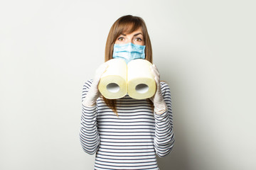 young girl in a medical mask and medical gloves, holds rolls of toilet paper, in the form of binoculars on a light background. Concept quarantine, shortage and panic