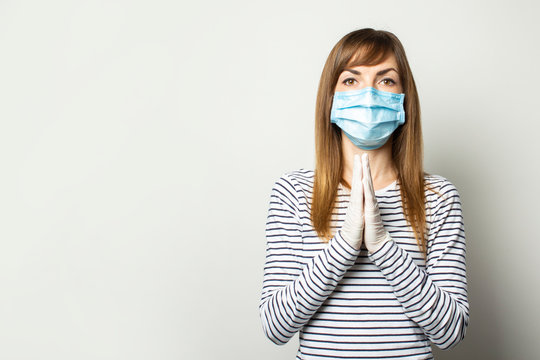 Young Woman In Protective Medical Mask And Latex Gloves Holds Her Hands Near Her Chest, Prayer Pose On A Light Isolated Background. Concept Of Coronavirus, Pandemic, Prayer, Appeal To God, Religion