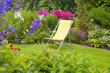 A beautiful blooming flower garden with yellow deck chair on a green lawn in summer
