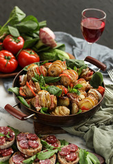 A summer lunch. Baked potatoes with bacon, cheese, tomato and herbs in a pan. A glass of red wine, bread with sausage, fresh tomatoes, cucumbers, garlic and spinach on the table. Background image