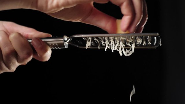 Professional chef rubs parmesan cheese on a grater. Grated cheese falls down on pizza with salami against black background. Concept of cooking delicious pizza. Slow motion. Close up