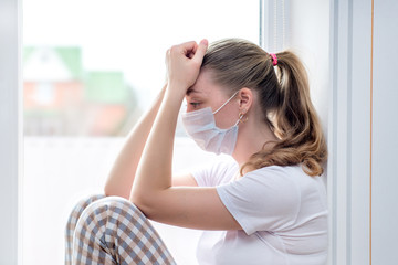 Home quarantine. Caucasian woman on sick leave sits at window in a medical mask, sad with her head in her hands. Poor health, a seasonal epidemic of the flu virus and colds. Home treatment