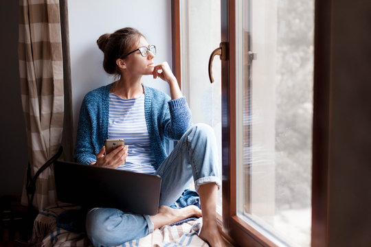Remote working from home office. Young woman using laptop, phone. Freelancer workplace by window. Teleworking in isolation, female business, shopping online, distance education. Lifestyle moment.