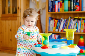 Happy joyful baby girl playing with different colorful toys at home. Adorable healthy toddler child having fun with playing alone. Active leisure indoors, nursery or playschool.
