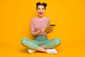 Obraz premium Full length photo of attractive lady sit floor legs crossed hold tablet open mouth read good news wear striped red white shirt green pants shoes isolated yellow color background