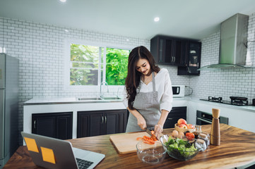 Image of young lady standing in kitchen using tablet computer and cooking vegetable salad with the tomatoes and avocado.