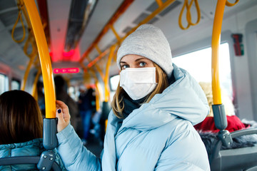 Blonde in medical mask standing in bus lounge next to yellow handrails in afternoon.