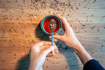 top view of female hands holding a teaspoon with tea leaves above a teacup on wooden table