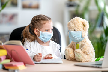 little girl in mask, with Teddy bear, doing homework, e-learning
