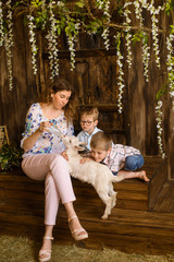 Mom with two sons sitting on wooden porch, watering white fluffy newborn goat from bottle with water