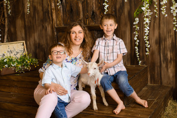 sitting young mother and two her sons kids with little white goat on dark wooden background with flowers and hay