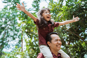 asian father carrying on neck smiling daughter while walking in the park - Powered by Adobe