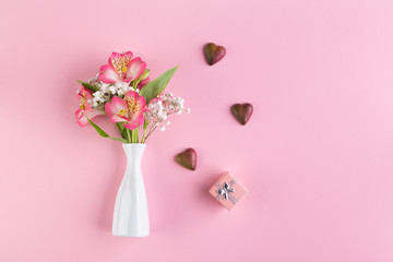 Alstroemeria and gypsophila flowers in a white vase, chocolate hearts and gift box on a pink background. Greeting card concept. Top view
