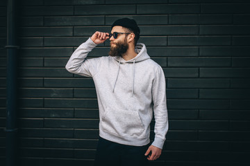 City portrait of handsome hipster guy with beard wearing gray blank hoodie or hoody and hat with...
