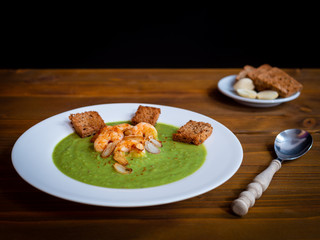 A plate of thick fresh green pea soup with prawns on a rustic wooden table with a garlic and croutons on the side.
