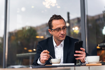 Businessman in outdoor cafe drinking cup of coffee while using mobile phone.