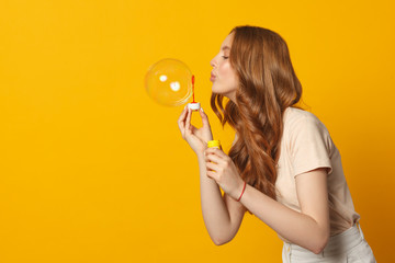 young female model blowing big soap bubble on yellow background, side view