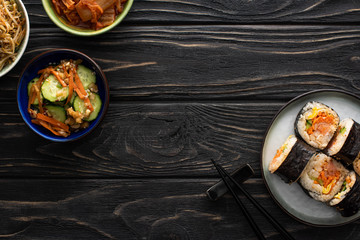 top view of plate with korean gimbap and chopsticks near tasty side dishes on wooden surface