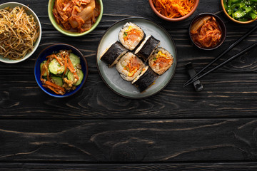 top view of plate with gimbap and chopsticks near tasty korean side dishes on wooden surface