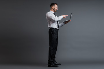 profile of angry businessman in suit pointing with finger at laptop on grey