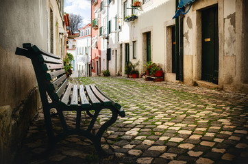 Fototapeta na wymiar Quiet narrow shady alley in the historical old district of Alfama in Lisbon, Portugal, with cobblestone and a wooden green bench in foreground and typical portuguese houses in background