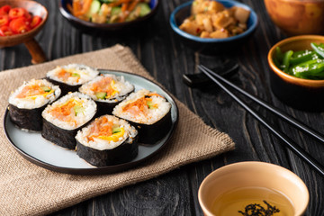 selective focus of tasty gimbap with vegetables and salmon near chopsticks and side dishes on wooden surface
