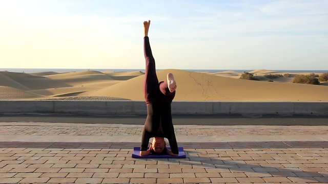 Female yogi practicing parivrtta eka pada sirsasana yoga pose by Maspalomas sand dunes. Flexible woman in revolved split-legged headstand asana. Challenge, balance, wellness concepts