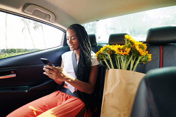 Beautiful positive young Black woman riding in taxi after supermarket and reading text messages...