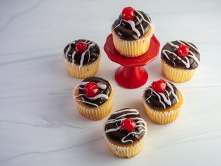 6, half dozen, yellow cupcakes with chocolate frosting and a white drizzle with a a red cherry on top and, sitting on a sandstone counter, with one cupcake raised up on a small red cupcake platter.