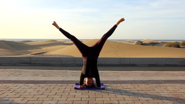 Female yogi practicing upavistha konasana in sirsasana at Maspalomas sand dunes. Iyengar yoga student doing headstand with open legs at natural environment. Balance, self care, flexibility concepts