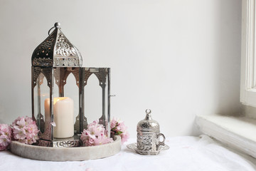 Silver decorative cup of tea, pink cherry tree blossoms and glowing Moroccan lantern on linen table...