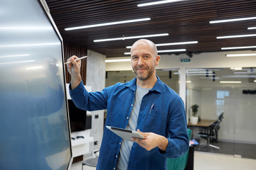 Waist up portrait of bald adult man smiling at camera while using digital board during presentation...