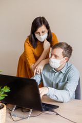 A young family works from home at the computer. Quarantined couple coronavirus in medical masks. The call to stay home safe. Order food products online. Laptop freelancer dispute business office