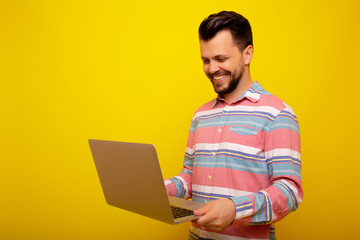 Portrait of happy smiling young man in casual wear, working with laptop, over yellow background. Image of caucasian man in striped shirt smiling while holding and using laptop. Copy space.