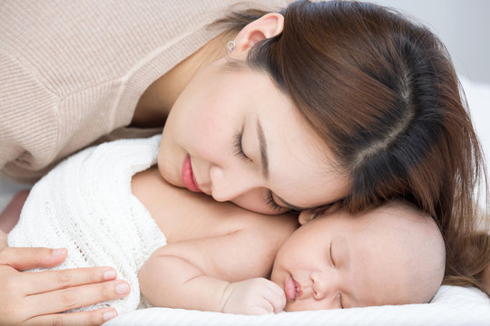 Beautiful Mom Supports And Tenderly Cuddles The Newborn Baby Gently While The Infant Is Sleeping On The White Bed. Asian Mother Using Her Cheek Touching On The Back Of Babies With Love And Care.