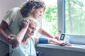 The girl assists an elderly woman in measuring blood pressure with a tonometer. Old people are exposed to the COVID-19 coronavirus. Daughter caring for a sick mother. A nurse helps a sick old woman