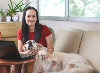 woman smiling and looking at  camera , sitting on couch in living room with computer in front of her and golden retriever dog lying on couch beside her.work from home concept