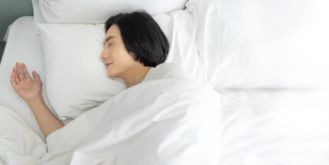 Fototapeta na wymiar Top view Young man sleeping and dreaming in white bed in the morning. Happy smiling Asian male enjoying lying on comfortable soft bedding in bedroom at home. eyes closed. copy space.