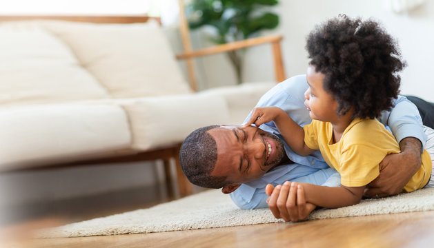 Happy African Father and little child son spending time playing at home together. Smiling Dad in blue shirt embracing or cuddling kid boy in yellow casual after arriving from working on wooden floor. 