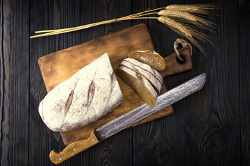 There is rye bread on the wooden village table . The bread is on an old cutting Board. Near the old big knife and ears of wheat. The view from the top