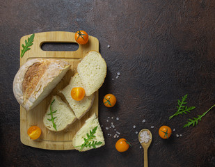 Rustic bread on wooden cutting board,yellow cherry tomatoes,frsh arugula leaves and pink sea salt on brown background.Top view.