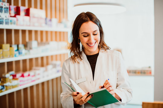 .Young female pharmacist working in her large pharmacy. Placing medications, taking inventory with her green notebook. Lifestyle