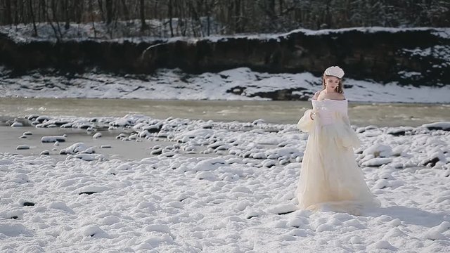 Amazing beautiful cute young woman in fairy tale image in white chic dress with crown stands on snow in sunny weather on background of mountain cold river