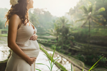 Young pregnant woman in white dress with view of Bali rice terraces in morning sunlight. View from...