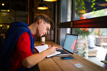 Young man college student writing in textbook during online education on laptop computer while...