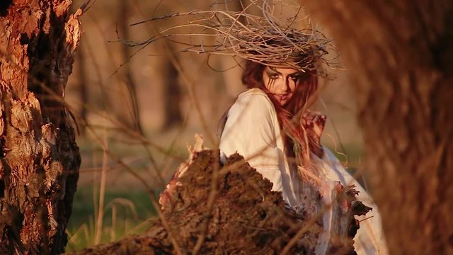 Young woman in form of witch sits before fire in forest among trees and conjures and dances on background of river
