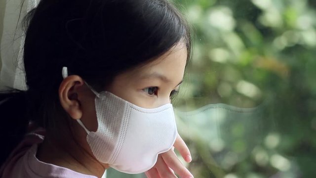 Asian 6 or 7 years old kid wearing medical mask.Little girl looking to outside through the window.She look sad,bored.She may sick or  individual quarantine from The Coronavirus Disease 19 (COVID-19).