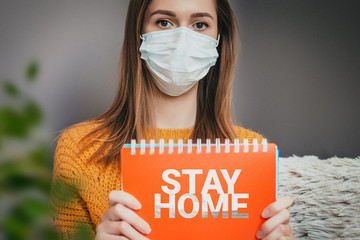 A young girl in a medical mask sits at home in isolation in quarantine, and holds a sign saying stay at home, coronavirus protection concept
